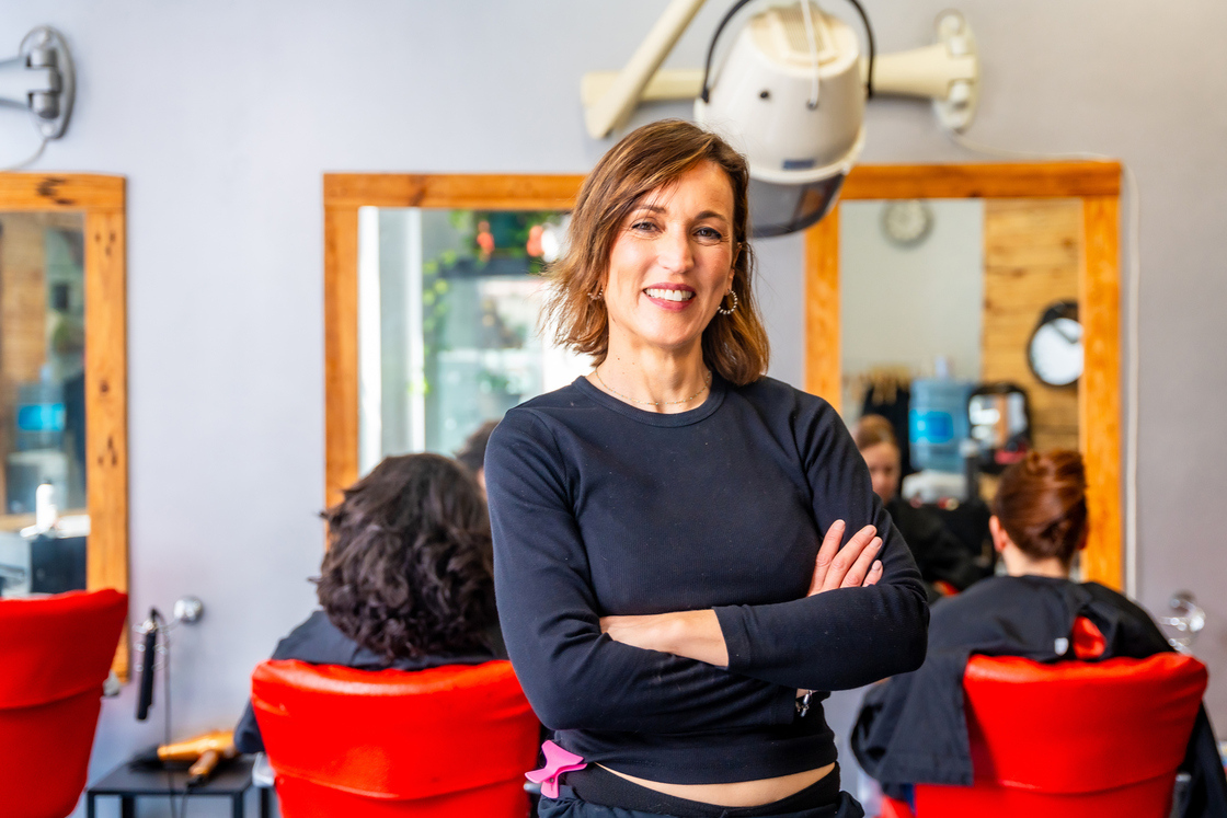 Imagem de uma cabeleireira sorridente em um salão de beleza, com cadeira vermelha e clientes ao fundo, transmitindo um ambiente acolhedor e profissional.