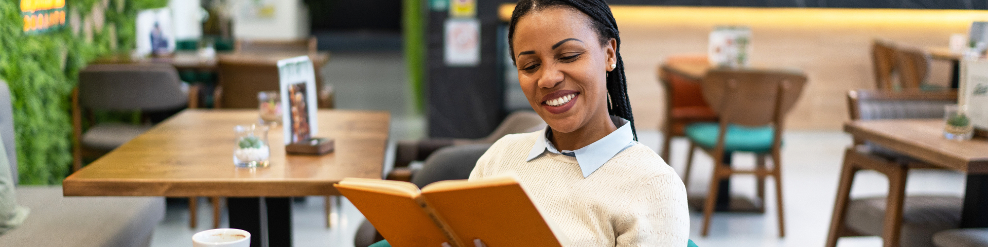 Mulher sorridente lendo um livro em um ambiente de café ou restaurante, com decoração moderna e ambiente aconchegante.