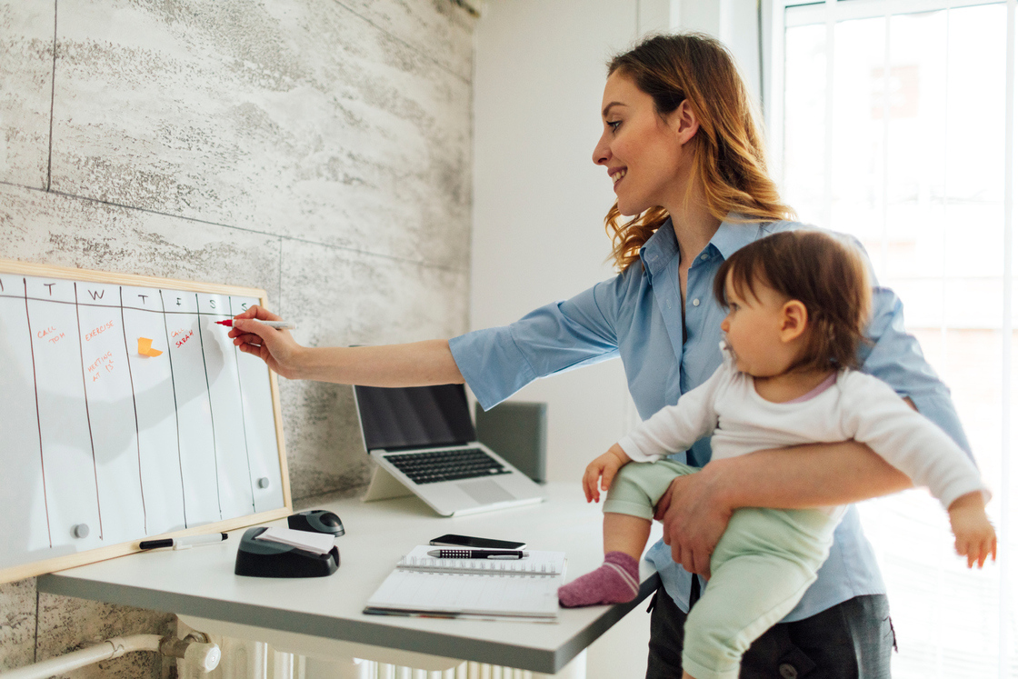 Mulher organizando o planejamento semanal enquanto segura uma criança em um ambiente de home office moderno e iluminado.