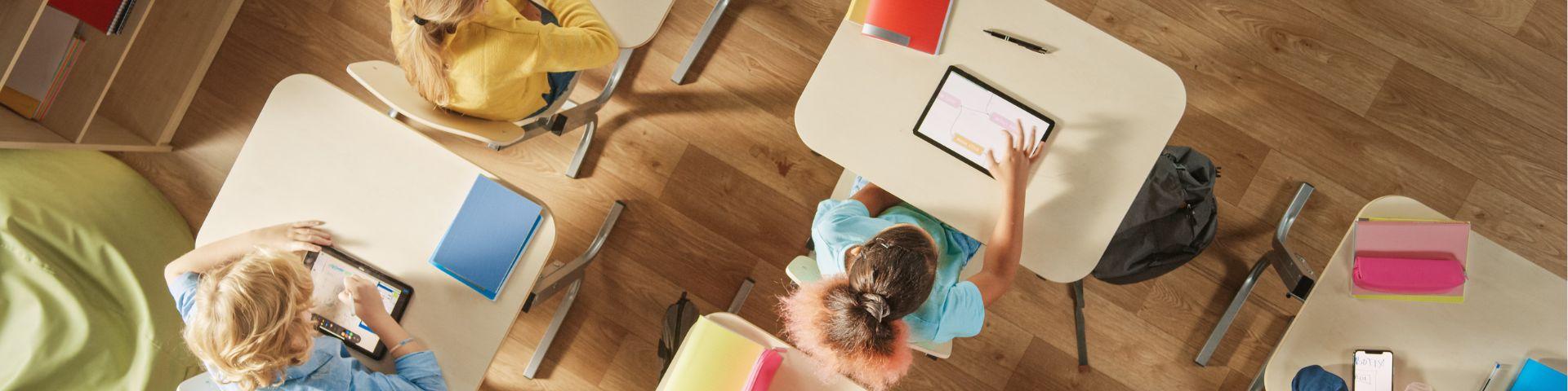 Sala de aula com crianças usando tablets e cadernos em suas mesas, ambiente escolar alegre e organizado.