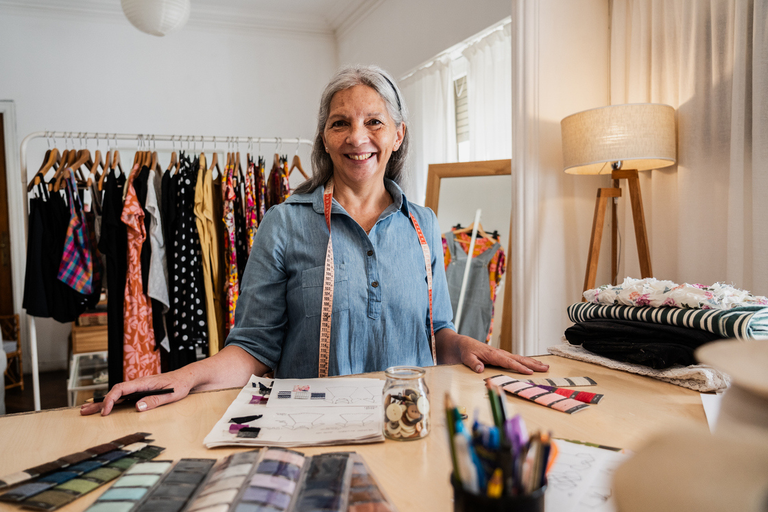 Mulher estilista sorrindo em sua loja, rodeada por roupas coloridas e materiais de costura, representando o mundo da moda e design. Com uma fita métrica ao redor do pescoço, ela inspira confiança e criatividade.