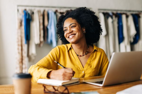 Mulher sorrindo enquanto escreve em um caderno, com laptop e café ao lado, em ambiente criativo e acolhedor. Ideal para ilustrar trabalho remoto e criatividade.