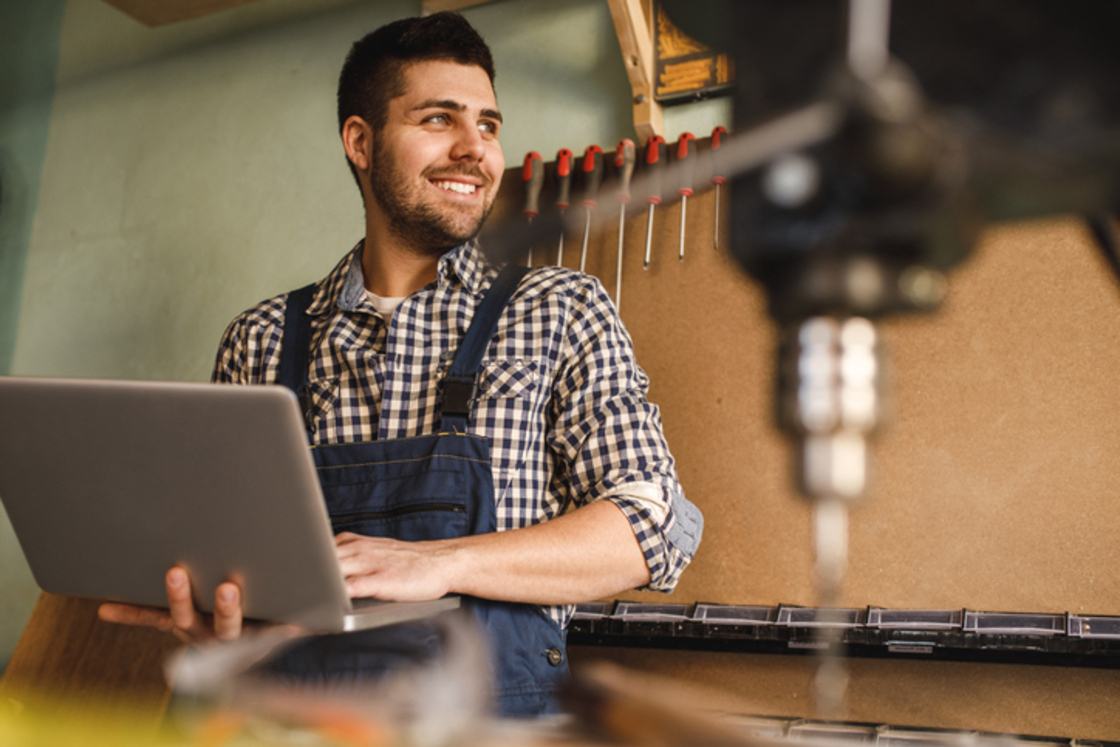 Homem sorridente usando laptop em ambiente de trabalho, rodeado por ferramentas, ideal para ilustração de trabalhos manuais e DIY.