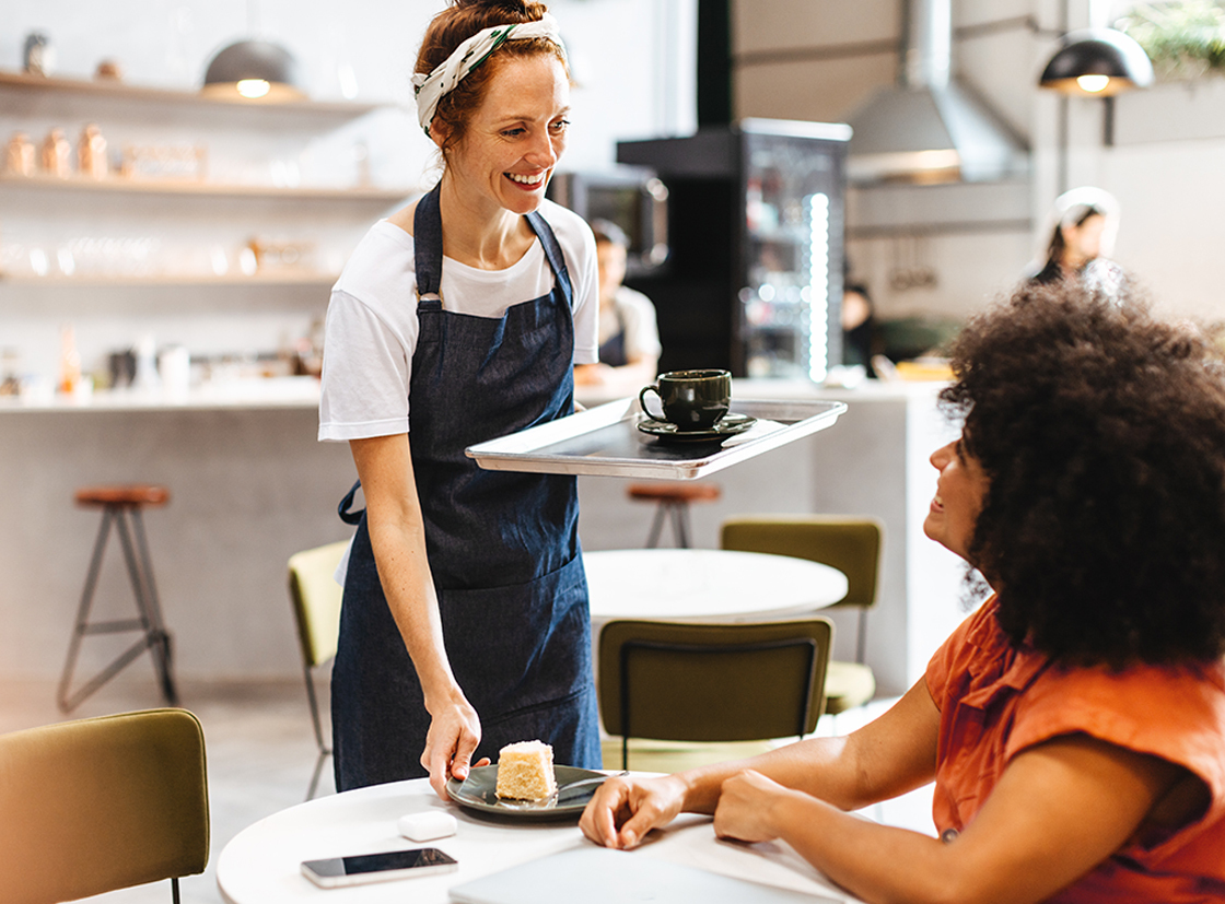 Uma garçonete sorridente serve café e sobremesa para uma cliente em um café moderno e aconchegante. O ambiente é iluminado e acolhedor, ideal para refeições.