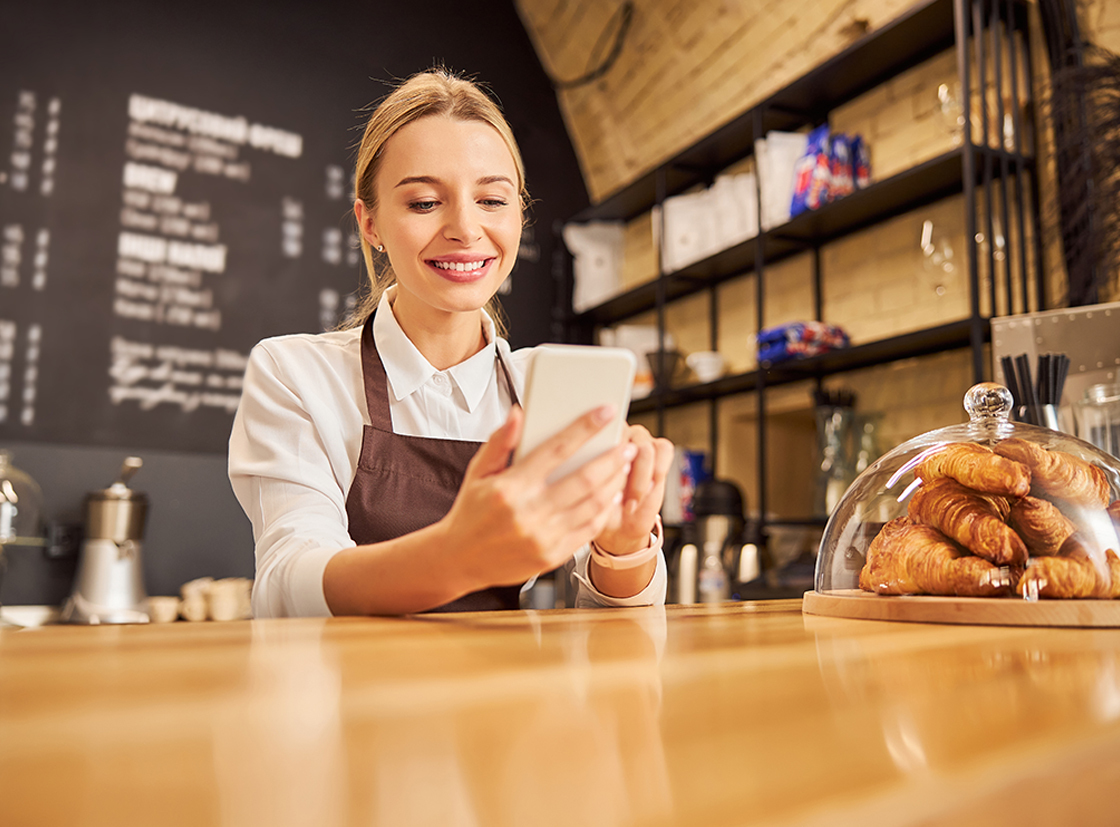 Mulher sorrindo em cafeteria enquanto usa smartphone. Ao fundo, croissants em uma vitrine. Atmosfera acolhedora e moderna.