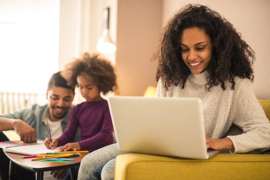 Uma mulher sorridente usa um laptop enquanto uma criança desenha ao seu lado, com um homem ao fundo, todos em um ambiente aconchegante.