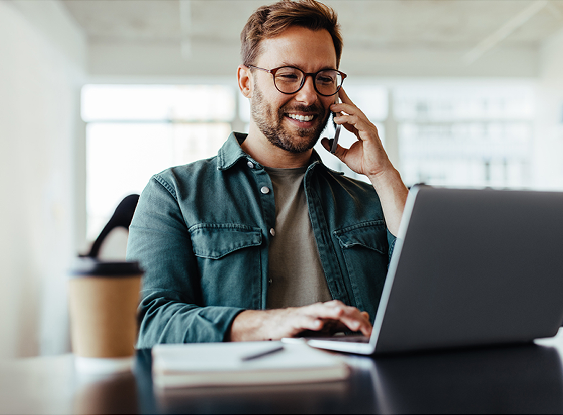 Homem sorridente em uma mesa usando laptop e falando ao telefone, com café ao lado, representando trabalho remoto eficiente.