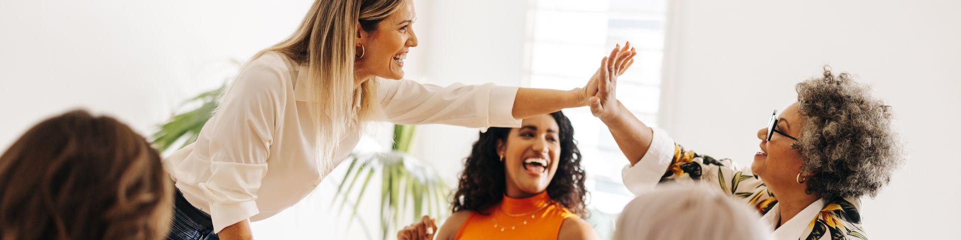 Grupo de mulheres sorrindo e interagindo em ambiente de escritório, demonstrando colaboração e bom humor.