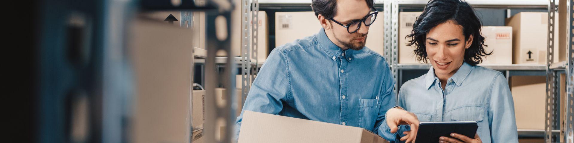 Dois empresários analisam produtos e discutem estratégias de logística em um armazém, simbolizando o planejamento e controle necessários para exportar com sucesso.