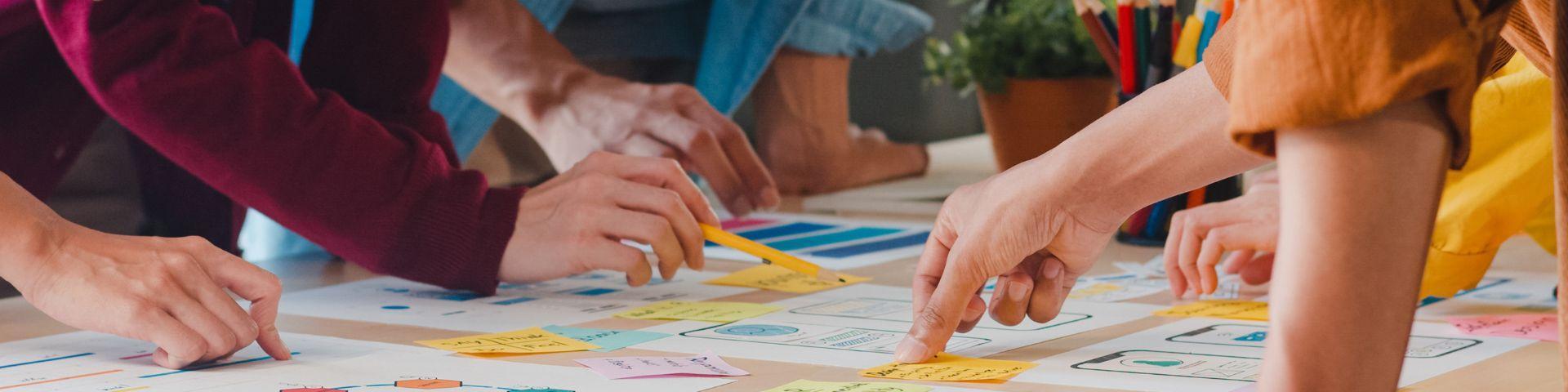 Equipe de colaboradores reunida em mesa de trabalho criativa com post-its, paleta de cores e lápis, representando práticas de inovação organizacional e metodologias colaborativas em pequenas empresas.