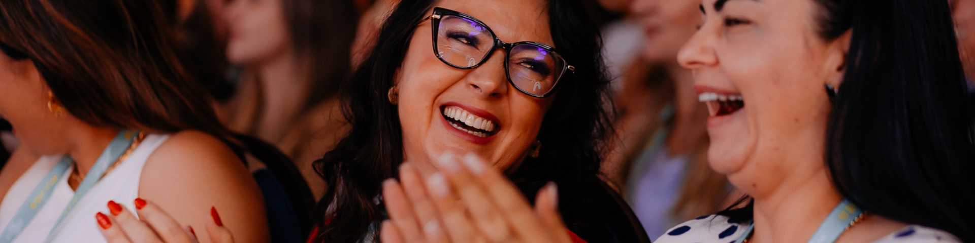 Mulheres sorrindo e rindo no Delas Summit, destacando-se a mulher de óculos de armação escura, usando vestido vermelho e acessórios dourados.