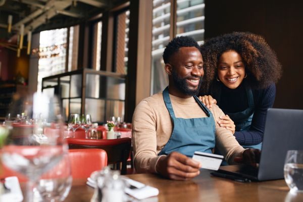 Um casal sorridente em um restaurante, verificando pedidos e pagamentos online com um cartão. A atmosfera é aconchegante e moderna, ideal para empreendedores.