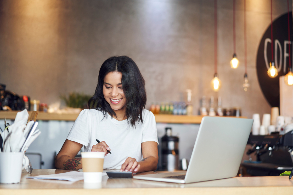 Jovem mulher sorridente, concentrada em seu trabalho em um café moderno, utilizando computador e calculadora enquanto toma café.