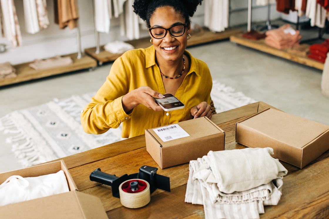 Mulher sorridente usando smartphone enquanto manuseia pacotes em uma loja, com produtos organizados ao redor.