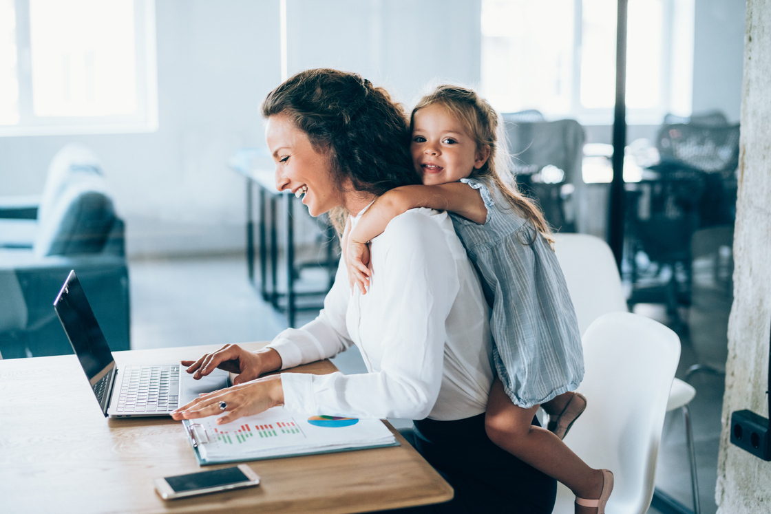 Mãe trabalhando em um laptop enquanto a filha se diverte nas costas dela, transmitindo a harmonia entre vida profissional e maternal.