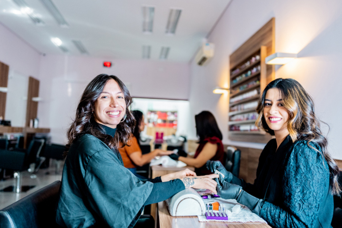 Duas mulheres sorrindo em um salão de beleza durante a manicure, com cores vibrantes e ambiente agradável.