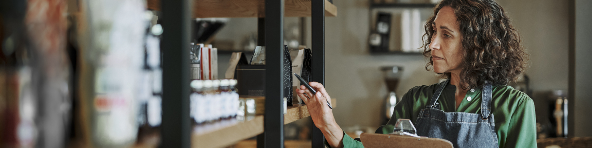 Mulher com cabelo cacheado usando uniforme de loja, verificando produtos na prateleira de uma loja de alimentos ou supermercado.