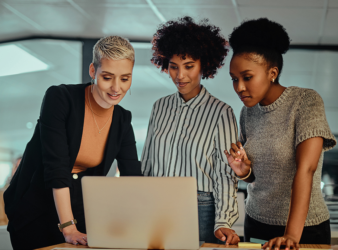 Três mulheres analisando um projeto em um laptop em um ambiente de trabalho colaborativo. Elas demonstram interesse e foco na discussão, representando diversidade e união.