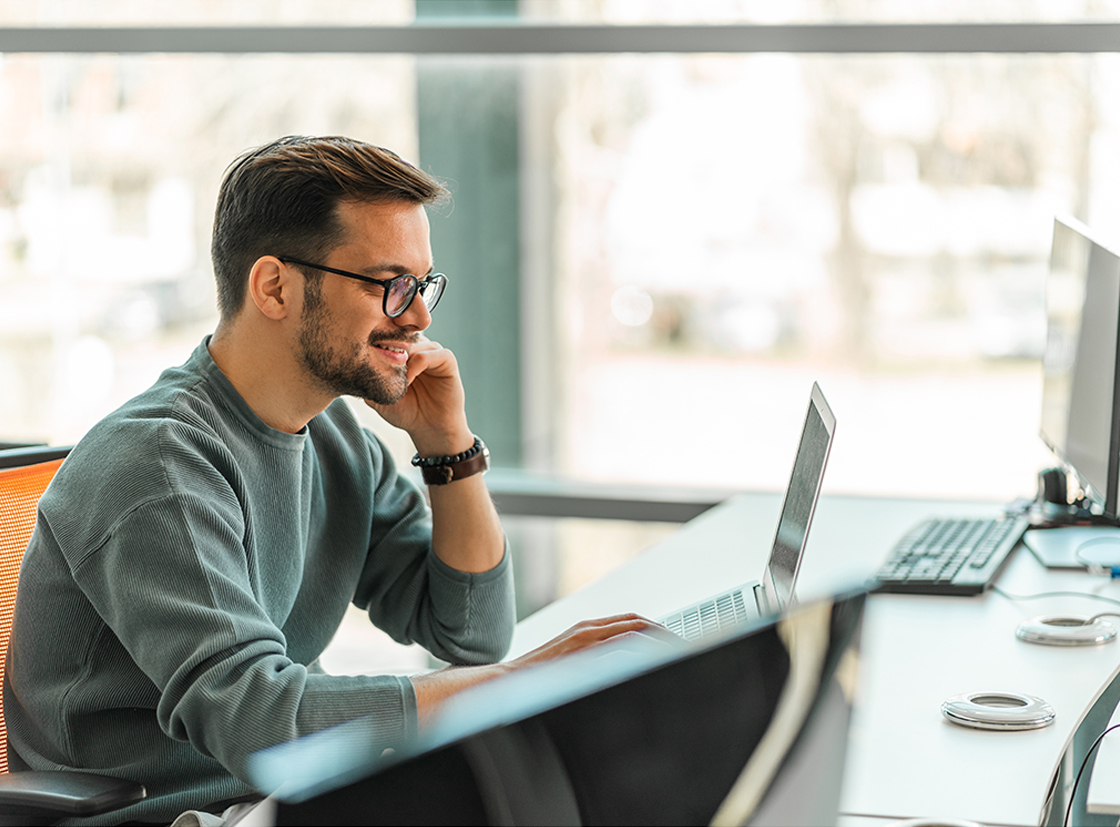 Homem de óculos, sorrindo e trabalhando em um laptop em um ambiente de escritório moderno, com mesas e computadores ao fundo.