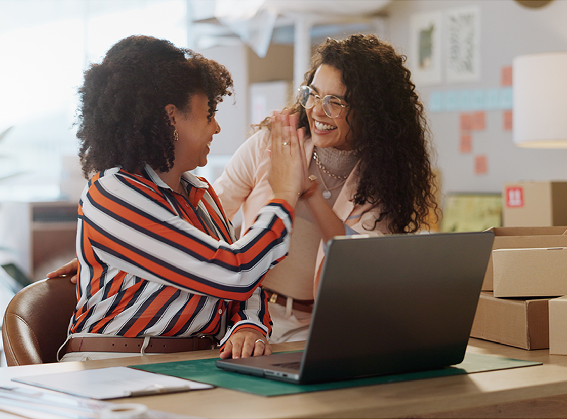 Duas mulheres sorrindo e se cumprimentando em um ambiente de escritório moderno com uma mesa e um laptop.