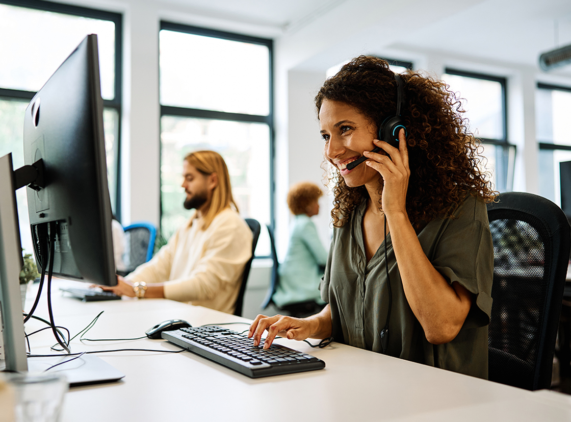 Mulher sorridente usando fones de ouvido enquanto trabalha em um escritório moderno, atendendo chamadas e interagindo com colegas.