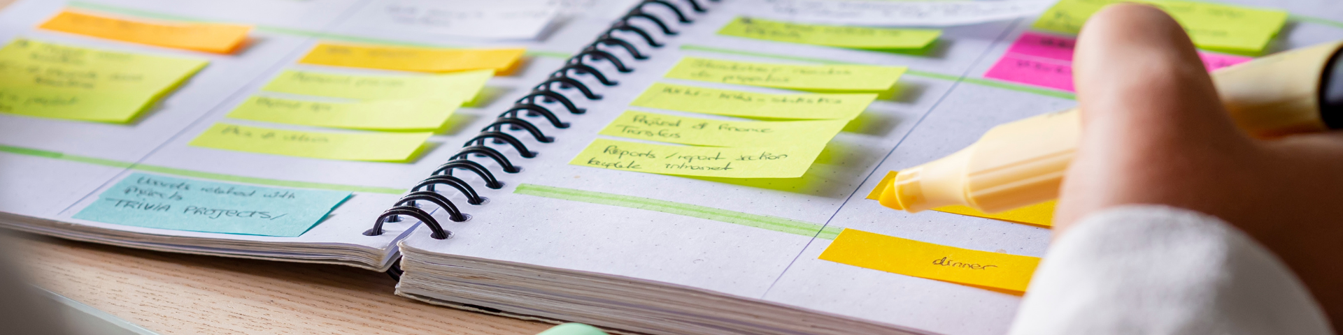 Pessoa organizando um planner com post-its coloridos em uma mesa de estudo, usando uma caneta para marcar tarefas, ideal para planejamento e produtividade.
