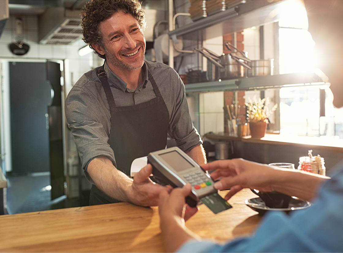 Homem sorridente atendendo um cliente em um café, utilizando um terminal de pagamento eletrônico. A cena retrata um ambiente acolhedor e amigável.