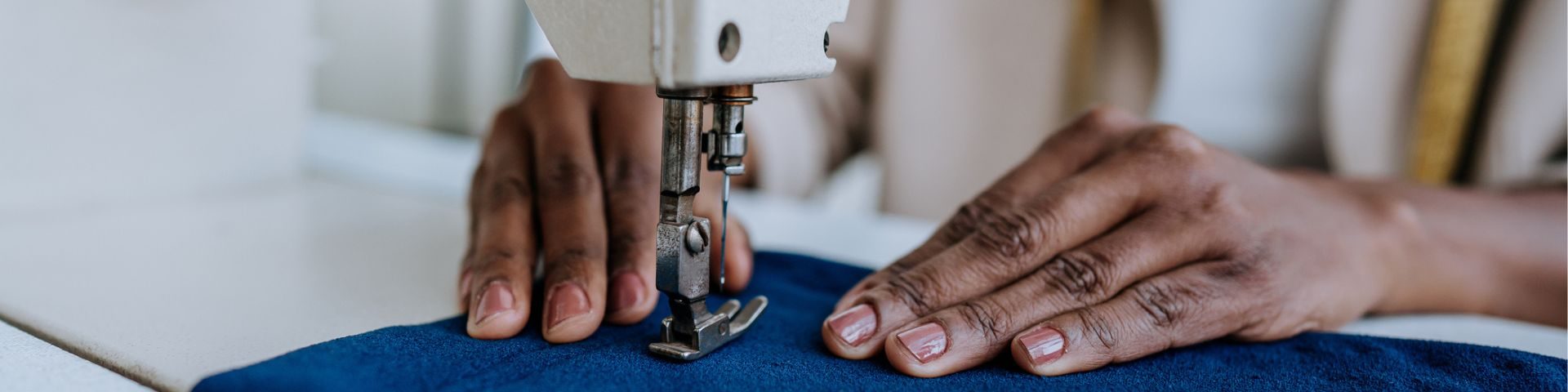 Pessoa usando máquina de costura para fazer roupas, foco na mão e na agulha em ação, ambiente de costura doméstica ou manufatura de roupas.