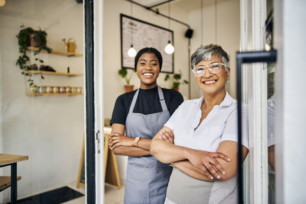 Duas mulheres sorridentes em um café, mostrando a alegria do trabalho em equipe e do atendimento ao cliente. Uma está vestida de uniforme, enquanto a outra usa roupas casuais, ambas com sorrisos acolhedores.