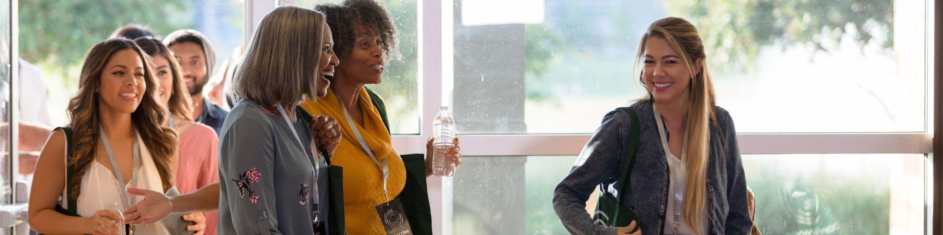 Grupo de mulheres empreendedoras chega a uma feira de negócios, trocando sorrisos e conversas animadas. A cena transmite entusiasmo, parceria e a importância da presença feminina no empreendedorismo.