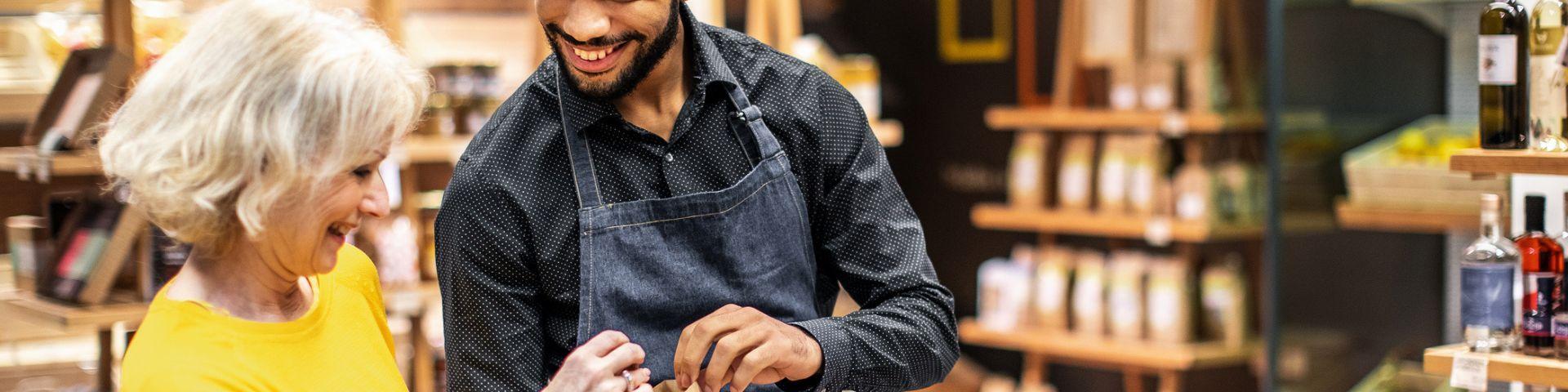 Homem ajudando idosa a escolher produtos em loja, ambiente de comércio com prateleiras de alimentos e bebidas ao fundo, sorriso no rosto.