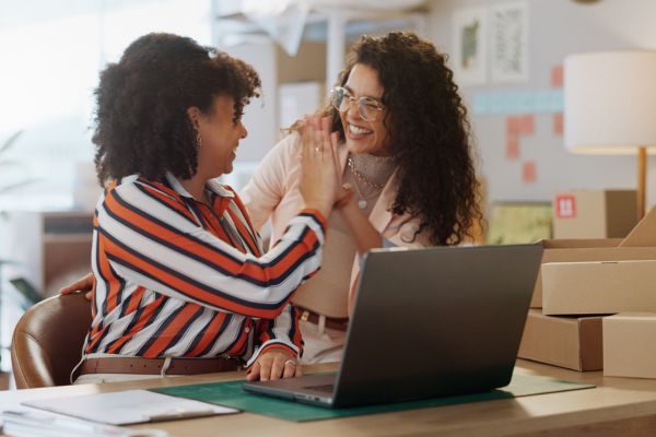 Duas mulheres sorrindo e se cumprimentando em um ambiente de trabalho, uma delas está usando uma blusa listrada e a outra com uma camisa clara, em frente a um laptop.