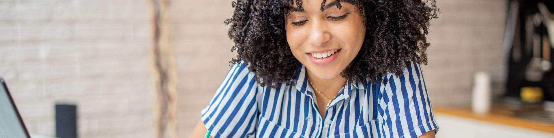 Jovem empreendedora sorridente fazendo anotações ao lado do notebook, ilustrando o planejamento e controle de resultados financeiros.