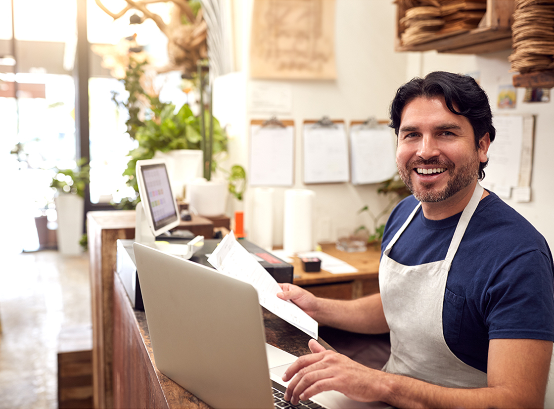 Homem sorridente em um café, trabalhando no computador e interagindo com clientes, mostrando o ambiente acolhedor do local.