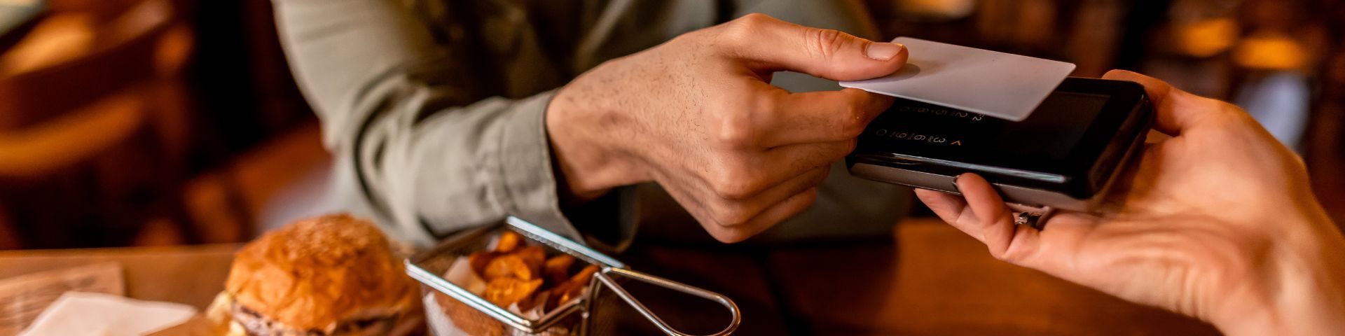 Pessoa passando cartão de crédito em uma maçaneta de mesa de restaurante, com comida ao fundo, em um ambiente de cafeteria ou restaurante sofisticado.