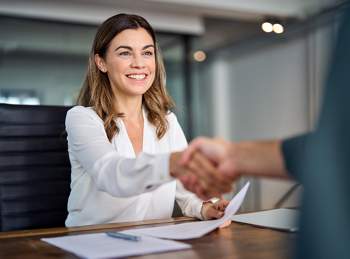 Mulher sorridente em ambiente de escritório cumprimentando com firmeza uma pessoa, simbolizando uma boa negociação e trabalho em equipe.