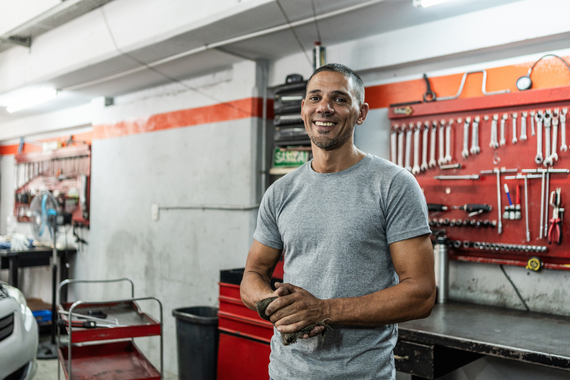 Homem sorridente em uma oficina mecânica, vestindo camiseta cinza, rodeado de ferramentas e equipamentos automotivos.