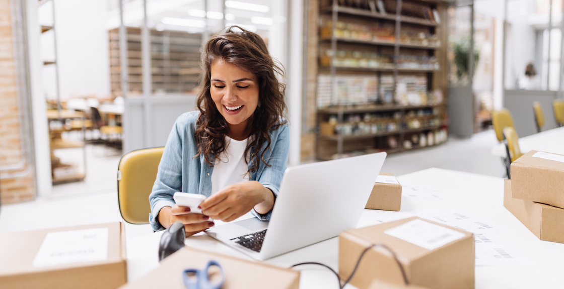 Mulher sorridente usando celular e laptop em ambiente de trabalho, rodeada por caixas e materiais de escritório, representando o empreendedorismo e a gestão de negócios.