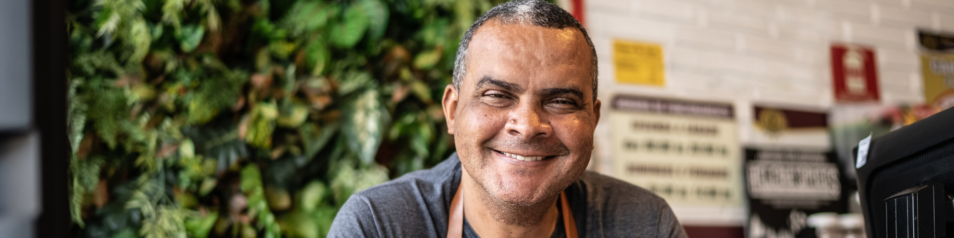 Homem sorridente em ambiente de loja ou mercado, com fundo de prateleiras e plantas ao fundo, transmitindo alegria e bom humor.