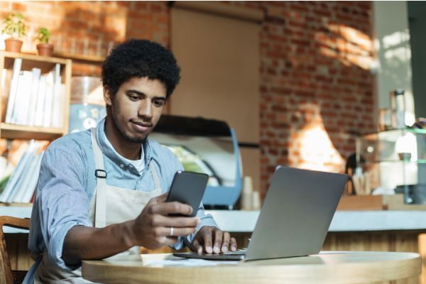 Homem jovem usando smartphone e laptop em um café, retratando um momento de trabalho moderno e prático.