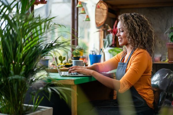 Mulher sorridente trabalhando em um laptop em um ambiente com plantas. Ela usa um avental e está concentrada em suas atividades, demonstrando produtividade e criatividade. Ideal para temáticas de trabalho remoto e estilo de vida saudável.