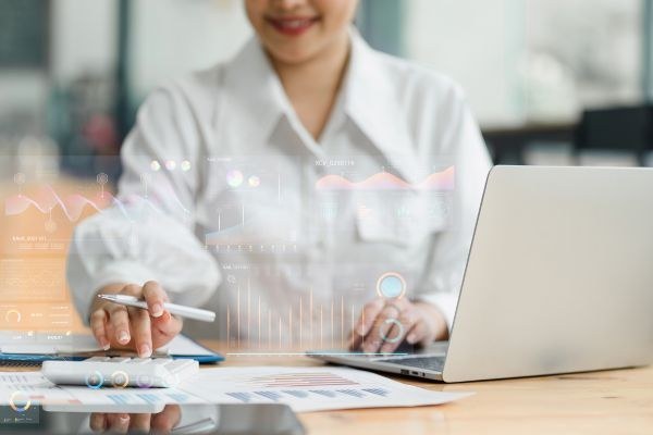 Mulher analisando gráficos e dados em um computador enquanto escreve em uma mesa. Ela está usando tecnologia para obter insights.