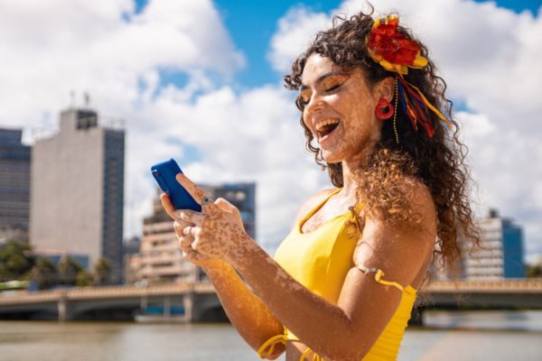 Mulher sorridente com cabelo encaracolado e flores, segurando um celular, em ambiente externo com prédios ao fundo. A imagem transmite alegria e conexão.