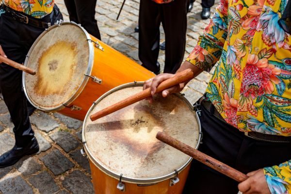 Imagens vibrantes de músicos tocando tambores em um evento cultural, com roupas coloridas, destacando a alegria da música ao vivo.