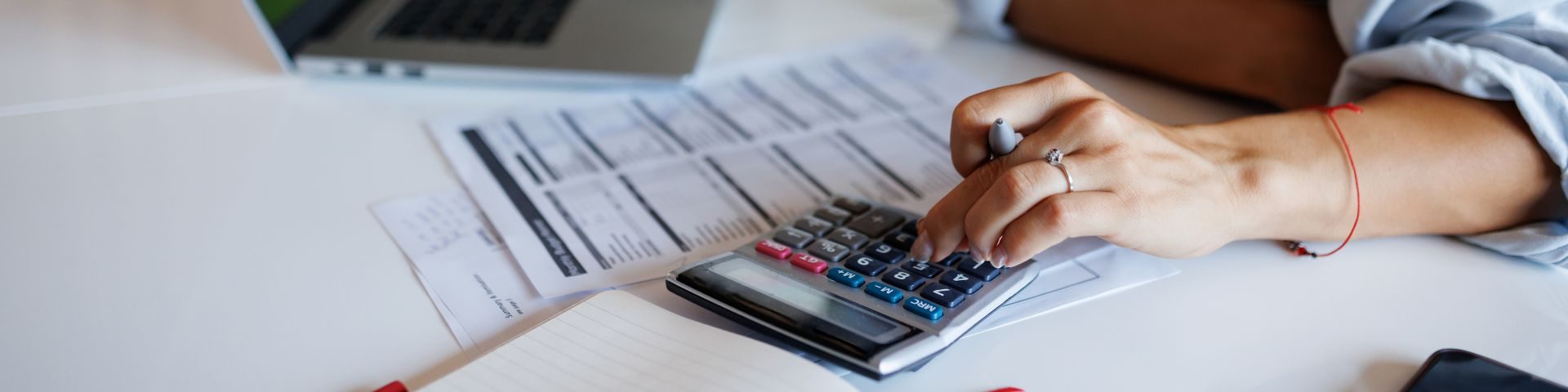 Jovem empreendedora branca vestindo camisa azul, calcula com auxílio de calculadora, planilhas em papel e notebook valores do INSS e aposentadoria privada para seu planejamento previdenciário.