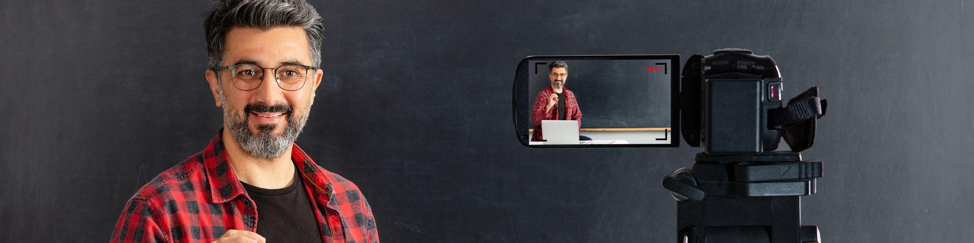 Professor apresentando conteúdo de frente para a câmera em um estúdio de gravação, com câmera filmando e quadro negro ao fundo.