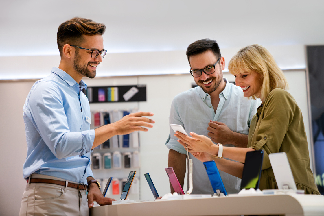 Grupo de amigos sorrindo enquanto escolhem smartphones em uma loja de eletrônicos, mostrando a interação social e a variedade de produtos disponíveis.