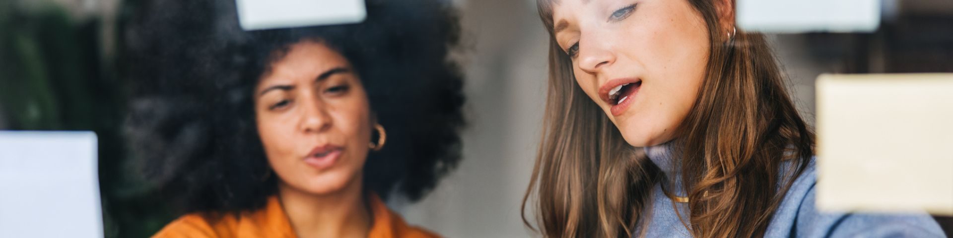 Duas mulheres discutem ideias em frente a uma parede de vidro com post-its coloridos. Uma delas, de cabelo cacheado e blusa laranja, gesticula enquanto fala; a outra, de cabelo liso e suéter azul, observa atenta e aponta para as anotações.