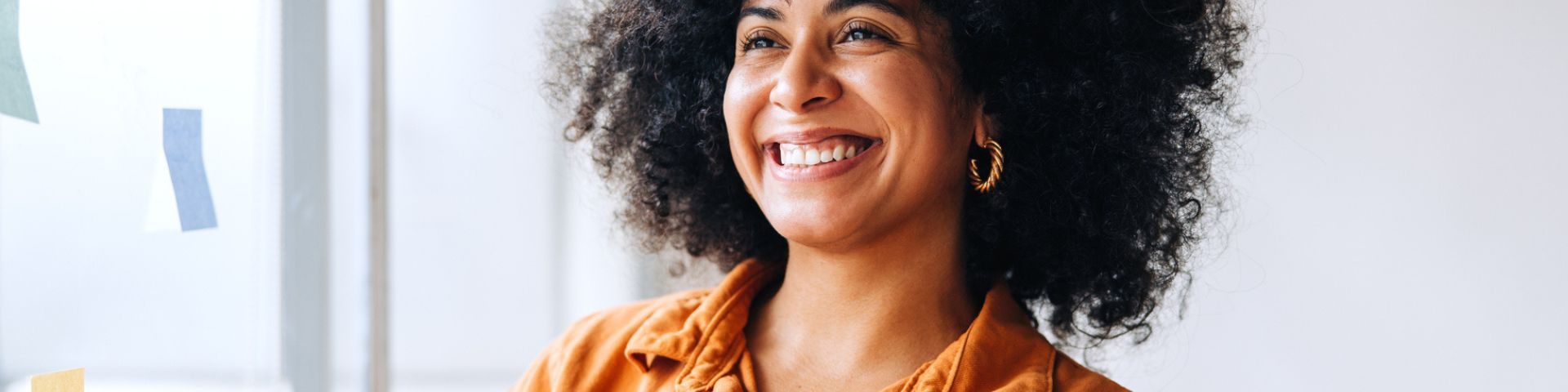 Mulher de cabelos cacheados e volumosos sorri, transmitindo leveza e confiança. Ela veste camisa laranja e brincos dourados, com fundo claro e post-its coloridos ao redor, em um ambiente de trabalho criativo e colaborativo.