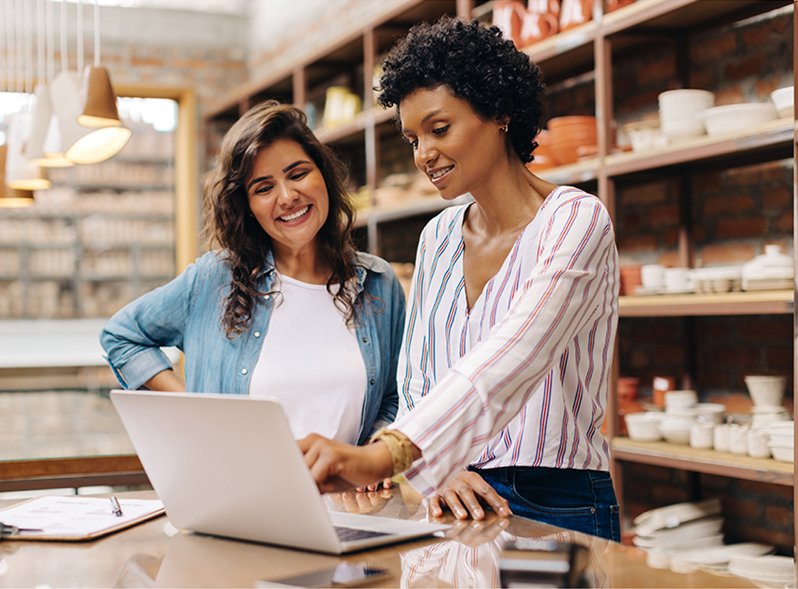 Duas mulheres sorrindo e interagindo enquanto trabalham em um laptop, em um ambiente agradável e iluminado. A cena sugere colaboração e criatividade.